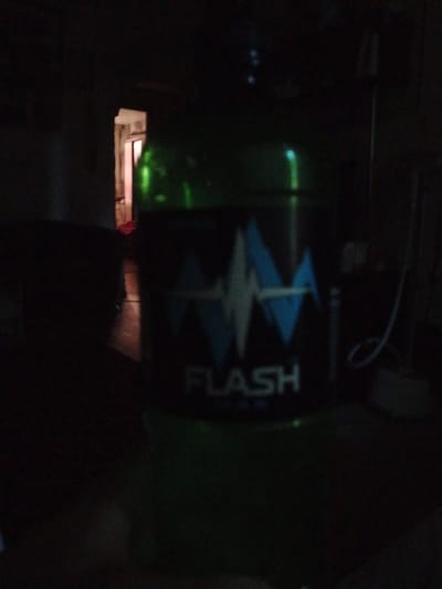 The image shows a dimly lit indoor scene. In the foreground, a green cylindrical object with a label is visible. The label has a graphic resembling an electrocardiogram or sound waves in blue and white, and the word "FLASH" in white capital letters. The background is dark, with a doorway visible in the upper left portion of the image, illuminated by a light source from beyond. The environment appears to be residential or commercial, but no specific details about the location in Guy, Russia, are discernable from the image itself. There are no people visible in the frame.