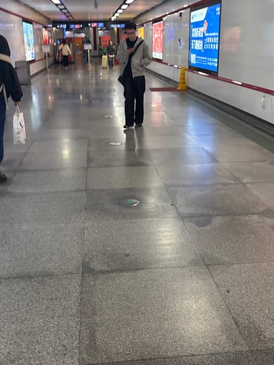 A gray tiled floor extends into the distance of an indoor space. A man in a gray jacket and black pants walks along the floor, with a white cone in the background. In the distance are various signs and posters, with a few other people visible. A person carrying a white shopping bag walks in the foreground.