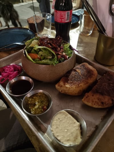 The image shows a food tray containing two pieces of bread, a salad in a brown bowl, and four small metal containers filled with dips. A bottle of Coca-Cola and a drinking glass are also visible. The location is likely in Mitzpe Ramon, Israel, given the visual style and the food served, which is consistent with Middle Eastern cuisine. No people are directly visible, but the presence of the food and drink suggests a dining setting. The environment appears to be indoors, possibly in a restaurant.