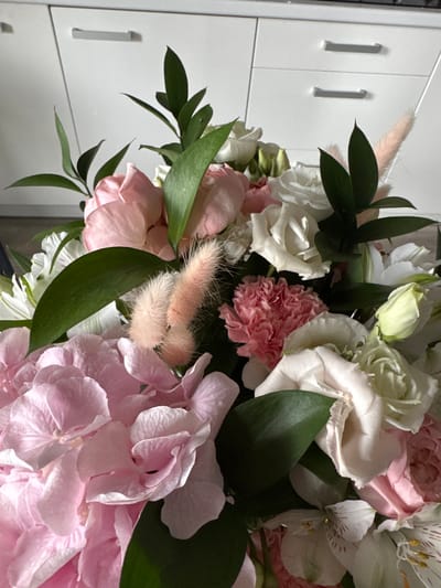The image shows a close-up of a bouquet of flowers, predominantly in shades of pink and white, with green foliage. The flowers include hydrangeas, roses, carnations, and alstroemerias. The bouquet is arranged in a dense, layered style. In the background, two white cabinet doors with horizontal silver handles are visible, suggesting an indoor setting, possibly a kitchen or living area. The lighting is bright, indicating daytime. The image does not contain any people or evidence of a specific location in Rzeszów, Poland.