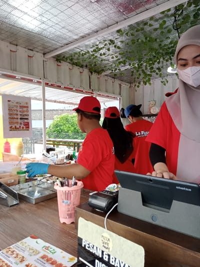 This bright, daytime image captures the interior of a casual food stall, likely a pizza shop, located in Kecamatan Panji, Indonesia. The setting appears to be a semi-open structure, possibly made from a converted shipping container or similar modular units, with a reflective, insulated ceiling adorned with artificial green vines.
The scene is bustling with several employees, all wearing matching red t-shirts, with "La Pizza" visible on some of their backs, and red baseball caps. In the foreground on the right, a person wearing a light grey hijab and a white face mask is seen operating a black tablet, which rests on a stand and has "sketsa" branding. This individual's hand is on the tablet, suggesting they are processing an order or payment.
To the left, another employee, wearing blue gloves, is actively engaged in food preparation on a stainless steel counter. Various ingredients, possibly pizza toppings, are laid out in trays, alongside condiment bottles and a shaker. A pink plastic caddy holding utensils sits on the wooden counter in front of the food prep area.
In the background, two more employees in red shirts and caps are partially visible, also attending to tasks. The overall impression is one of an active, efficient food service operation.
On the wooden counter in the lower left, a printed menu displays pizza options, with visible text including "CLASSIC CHEESE 17K" and "MEAT LOVERS 26K", along with "PEPPERONI." A prominent sign in the foreground clearly states "PIZZA TAKE AWAY" and "PESAN & BAYAR DI SINI," which translates to "Order & Pay Here," indicating the shop's service model. Through the open sections of the stall, bright daylight and green trees are visible, suggesting an outdoor or semi-outdoor dining area beyond the counter.