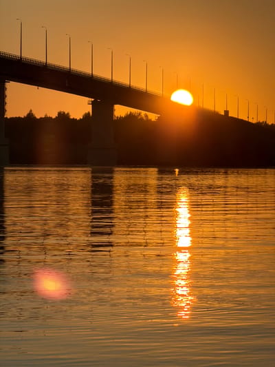 This is a photograph of the sun setting over the Kama River in Kambarka, Russia. A large concrete bridge with streetlights spans across the frame. The sun is partially visible behind the bridge, casting a warm, orange glow across the sky and reflecting on the rippling water. Trees and their silhouettes are visible along the riverbank. The scene is devoid of people.