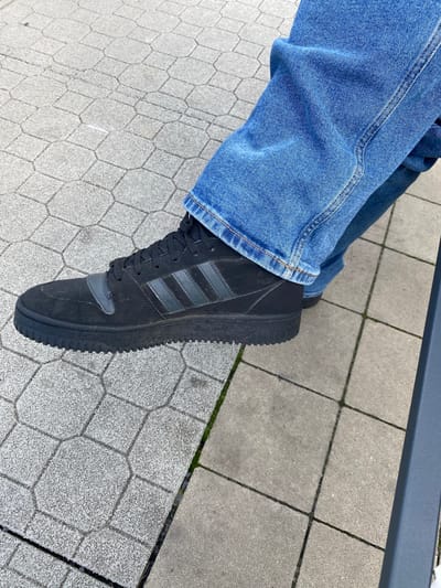 The image shows a close-up, ground-level view of a person's left foot wearing a black Adidas sneaker, with blue denim jeans covering the ankle and lower leg. The sneaker has dark gray Adidas stripes on the side and a dark gray strap across the front. The person is standing on a textured, light gray paver walkway with a repeating hexagonal pattern, typical of public outdoor spaces. The walkway appears to be in good condition. The scene is set outdoors, with the lighting suggesting it is daytime. No other people or specific landmarks are visible, making it difficult to pinpoint a precise location within Lahošť, Czechia, beyond the general context of an outdoor paved area.