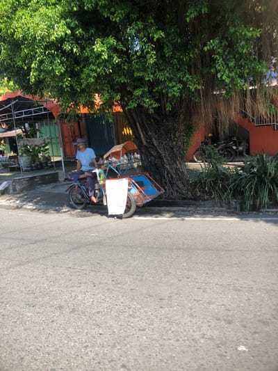 A man wearing a light blue t-shirt, dark pants, and a gray cap is seated on a motorbike that is attached to a pedicab. The man has his left leg crossed over his right, with his bare feet visible. He is wearing a dark face mask. The pedicab is blue with red accents and has an orange canopy. A white cloth is attached to the front of the pedicab. The motorbike and pedicab are parked on the side of a paved road, partially shaded by a large tree with thick, gnarled roots and hanging aerial roots. In the background, there are buildings with orange and green facades, as well as some potted plants and what appears to be bottled water stacked up. Sunlight is casting shadows across the scene, indicating it is daytime. The scene is located in Buduran, Indonesia.
