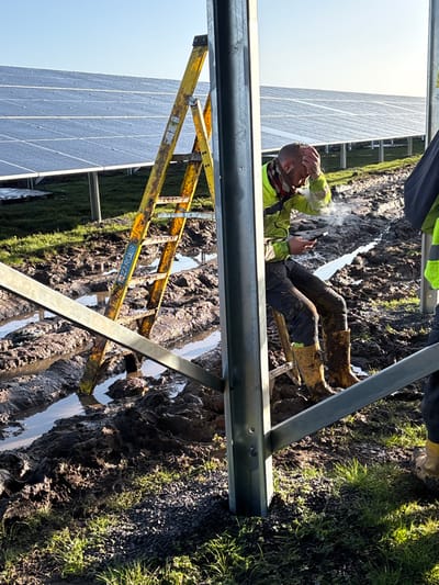 The image depicts an outdoor construction site within a solar farm in Grange Ville, Ireland, under clear daylight conditions. The ground is heavily disturbed, characterized by deep, wet mud with standing water collected in ruts and depressions, indicative of recent vehicle activity or precipitation. Patches of green grass and gravel are visible at the immediate foreground.

In the mid-ground, a male individual is seated or squatting on a mound of mud, facing towards the right side of the frame. He is dressed in work attire consisting of a neon yellow high-visibility jacket, dark trousers, and mud-splattered yellow rubber boots. A patterned scarf or snood is worn around his neck. His right hand is raised to his forehead, while his left hand holds a black smartphone. Visible vapor emanates from his mouth area, suggesting exhalation in cold air. His expression appears strained or contemplative.

To the left of this individual, a yellow metal A-frame stepladder, bearing the brand "ZARGES," stands partially submerged in the mud and water. The ladder appears weathered and dirty. In the background, rows of installed solar panels extend across the field, with their surfaces appearing wet or reflective.

Prominently in the foreground and mid-ground are galvanized steel structural elements, likely supporting the solar panels, with diagonal bracing visible. A portion of a second person, wearing similar high-visibility workwear and yellow rubber boots, is partially visible on the far right of the frame. The scene conveys a challenging work environment for solar panel installation.