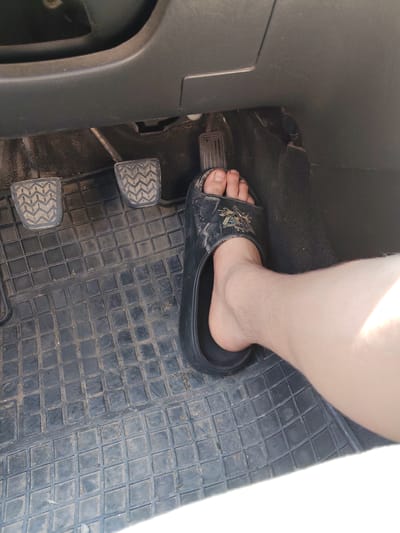 The image shows a close-up view of a person's foot wearing a black slide sandal, positioned on the floor of a car. The foot is on the gas pedal, which is visible on the right side of the frame. To the left of the gas pedal are the clutch and brake pedals, also visible, and the car's floor mat. The floor mat is dark gray with a textured, grid-like pattern and appears to have some dust and debris on it. The car's interior dashboard and lower console are visible in the background, in dark gray plastic. The lighting suggests it is daytime, likely sunny, as indicated by the bright highlight on the person's leg. The overall scene is a mundane view of the driver's side footwell of a vehicle, with no specific text visible in the image. The context of Ukraine, Moldova is provided but not directly evident from the image itself.