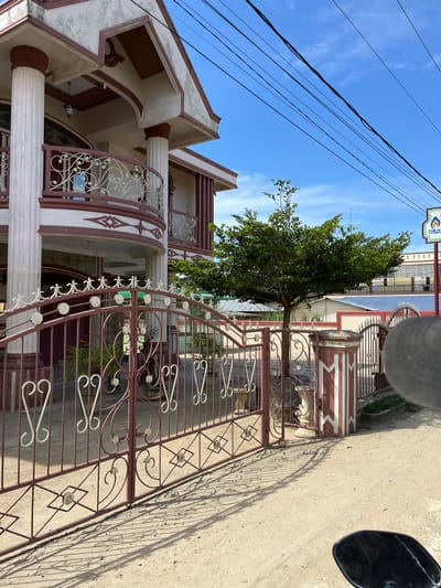 The image captures a street-level view in the city of Dalam, Indonesia. A large, two-story house with prominent columns and a decorative balcony dominates the left side of the frame. The house is painted in cream and burgundy colors. In front of the house is a large, ornate burgundy gate with intricate metalwork. Beyond the gate, a leafy green tree stands in the yard. The ground is a dirt path with shadows cast by the gate and tree. In the background, there are other buildings and a clear blue sky with power lines strung across it. A sign with a logo that appears to read "ELPIJI" is visible on the right side of the image. The perspective suggests the photo was taken from a vehicle, possibly a motorcycle, as the edge of a black mirror is visible in the lower right corner.