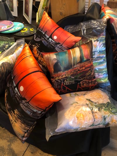 The image shows a close-up of a table display featuring several decorative pillows and round decorative items. The pillows have vibrant, artistic designs. One pillow in the foreground features an orange and black abstract pattern that resembles a sunset over water with boats. Another pillow to the right displays a market scene with stalls of fruits and vegetables, and a person in the background. A third pillow, visible in the lower right, is covered in plastic and has a floral print of light-colored roses. Two round items are stacked on the left side of the image; the top one is purple with "Welcome" written on it and decorated with flowers, and the bottom one is teal and black with a sunflower design. The items are laid out on a black fabric surface, with some wooden planks visible below. In the background, there are framed artworks and what appear to be shopping bags. The lighting suggests an indoor setting, possibly a market stall or craft fair. The city of Severn, Canada, is not depicted or suggested in the image.
