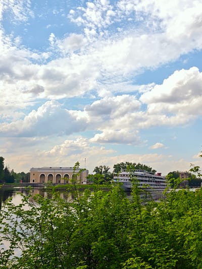The image shows a landscape with a river or canal, a building, a cruise ship, and lush greenery. The building has a neoclassical architectural style with a series of arched openings. The cruise ship is multi-tiered and appears to be docked or moving slowly on the water. The sky is filled with puffy white clouds and patches of blue. The scene suggests a tranquil setting, possibly in a recreational area of Водник Снт, Russia. The foreground is dominated by green foliage, partially obscuring the view of the water and the distant objects.