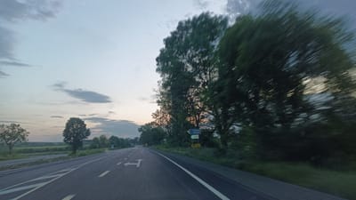 The image shows a two-lane road with white directional markings. The sky is overcast with some clouds. On the left side of the road is a grassy area with trees. On the right side, there are trees and a road sign indicating directions to "Sokolovo 1 km" and "Zlatintsa". The image appears to be taken from a moving vehicle, as some of the trees on the right side of the road are blurred. The overall scene suggests a rural or semi-rural environment, likely a road approaching or leaving the city of Sokolovo, Bulgaria.