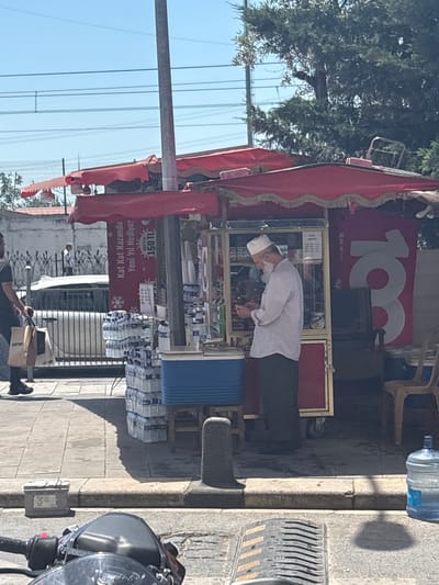 This image depicts a bustling outdoor street scene in an urban area of Gaziosmanpaşa, Turkey, under bright daylight with a clear blue sky. The central focus is a red street kiosk, covered by a large red canopy with gold-fringed trim. The kiosk features a glass display case on its left side and a solid red panel on its right. A red banner with white Turkish text is affixed to the front, reading "Kat Kat Kazandıran Yeni Yıl Hediyesi" (Winning Multiple New Year Gifts) and displaying "150 TL" along with a snowflake graphic. Another red banner on the right side of the kiosk shows a large white number "103". Standing behind the kiosk, on the left side, is an older man with a white beard and a white skullcap, wearing a light-colored long-sleeve shirt and dark pants. He is looking down at a small object held in his hands. To his left, on the sidewalk, is a blue insulated cooler, and stacks of bundled small plastic water bottles are arranged on the ground and on a metal stand. More large plastic water bottles are visible to the right of the kiosk. Two brown plastic chairs are situated on the sidewalk to the far right of the kiosk. On the left side of the image, a man wearing a dark short-sleeve shirt, dark shorts, and dark sandals is walking away from the kiosk towards the left, carrying two paper bags in his left hand, one visibly branded "KOTON". In the background, behind a decorative black metal fence, a white car is visible on the street. A distant, lightly dressed figure can be seen walking behind the car. Overhead, multiple power lines traverse the blue sky. A tall, gray utility pole stands between the two red banners of the kiosk. Green trees and foliage are visible in the background, particularly on the right side. The foreground shows parts of a dark-colored motorcycle or scooter, including the handlebars and front panel, indicating the perspective of the photograph. The sidewalk is paved with concrete slabs, and a gray concrete curb separates it from the asphalt street. A small, square, gray utility box rests on the sidewalk near the curb. In the immediate foreground, a gray metal object with horizontal ridges, possibly a speed bump or drainage cover, is on the road surface. Shadows from the kiosk and various objects are distinctly cast on the sunny