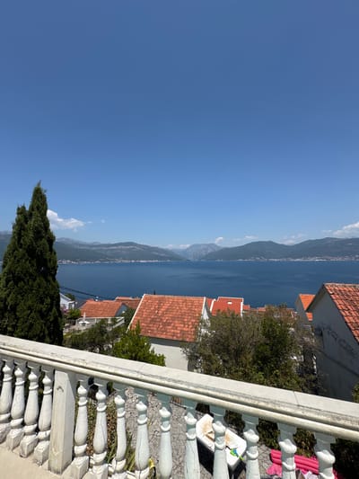 The image captures a scenic view overlooking the Bay of Kotor from Krašići, Montenegro. In the foreground, a white balustrade with ornate carvings separates the viewer from the scene below. Beyond the balustrade, several houses with characteristic terracotta roofs are nestled amongst lush green trees. In the middle ground, the vast expanse of the deep blue Bay of Kotor stretches out, bordered by rolling, verdant mountains that rise majestically in the distance under a clear blue sky. The overall impression is one of tranquility and natural beauty.