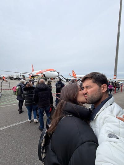 The image depicts an airport tarmac in Colombier-Saugnieu, France, likely Lyon-Saint-Exupéry Airport (LYS), under an overcast sky. In the immediate foreground, two individuals, a female with long brown hair wearing a black puffer jacket and a male with short brown hair and stubble wearing a white puffer jacket, are positioned right of center. They are engaged in a kiss, with the male's eyes visibly squeezed shut and his lips pursed.
Behind the couple, a queue of approximately 15-20 passengers is visible, standing on an asphalt tarmac marked with white lines and delineated by red and white striped barrier tape on the left. In the midground, two orange and white EasyJet aircraft are parked. The prominent aircraft, an Airbus A320 type, is being boarded by passengers via a mobile airstair at its front left door. A ground service vehicle and a ground crew member in an orange high-visibility vest are positioned near this aircraft's nose. A second EasyJet aircraft is partially visible further back on the right. A tall, slender grey pole stands vertically on the far right side of the frame.