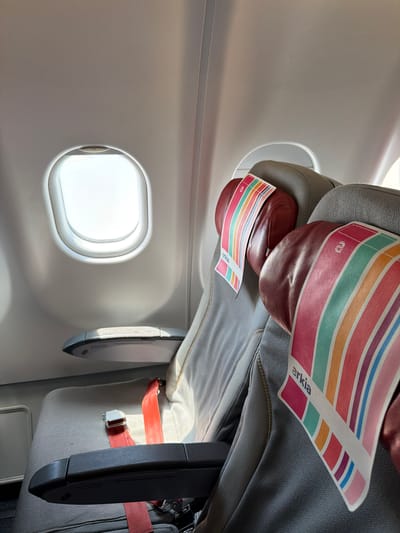 The image shows the interior of an airplane cabin. A window is visible on the left side of the frame, allowing bright light to enter. An airplane seat is present, with an "arkia" branded headrest cover. A seatbelt and a portion of a suitcase are also visible. There is no indication of the image's location being in Tzafria, Israel.