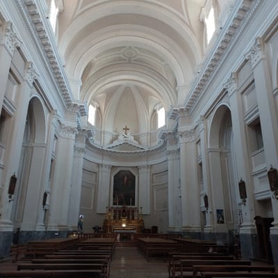 The image displays the interior of the Chiesa di San Domenico in Ancona, Italy. The view is centered on the nave, extending towards the main altar and dome. The architecture features a high, vaulted ceiling and a series of light-colored, fluted columns with ornate capitals supporting arches and a prominent entablature along both sides of the nave. Natural light illuminates the upper sections of the church through numerous high-set windows along the nave and at the base of the central dome.

The chancel area, elevated from the nave, prominently features a large, dark-toned religious painting positioned above an ornate golden altar table. A cross is centrally mounted atop a classical pediment above the painting. Rows of vacant wooden pews are systematically arranged on a darker-toned floor, forming a central aisle leading directly to the altar.

One individual is partially visible, seated in a pew on the far right side of the image. No other persons are discernible within the scene. Several rectangular religious artworks are mounted vertically on the columns along the side aisles. A statue, partially visible on the left, appears to be clothed in blue. The overall environment is quiet and spacious.