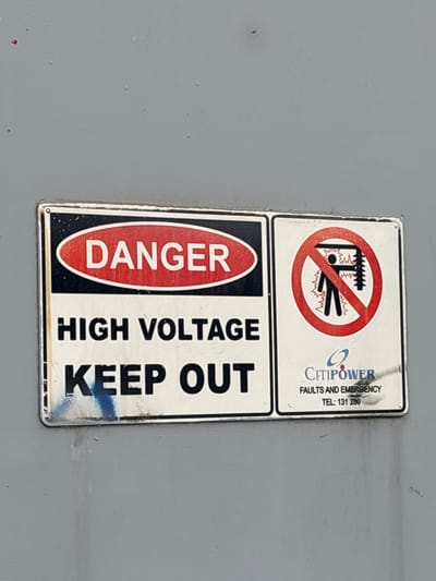 A close-up, eye-level, medium shot captures a rectangular sign with a white background and black border affixed to a light gray metallic surface. The sign is divided into two sections. The left section features a red oval with the word "DANGER" in white capital letters, positioned above the words "HIGH VOLTAGE" and "KEEP OUT," all in black capital letters. The right section displays a circular red prohibition symbol over a black silhouette of a person standing next to a power line, with zig-zag lines indicating electricity. Below this graphic, the text "CITIPOWER" appears in blue, followed by "FAULTS AND EMERGENCY" and a telephone number "TEL: 131 280." The metallic surface shows minor scuffs and a faint blue streak across the bottom of the sign. The scene is located in Brunswick East, Australia.
