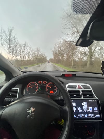 The image depicts the interior view from the driver's seat of a Peugeot vehicle on a wet, overcast day in Topolovgrad, Bulgaria. The road, a two-lane asphalt surface, is visible through the windshield, appearing wet from rain or damp conditions. Bare deciduous trees line both sides of the road, forming a dense backdrop against a uniformly gray, overcast sky. A red vehicle is observed in the distance, proceeding in the same direction on the road.
Inside the vehicle, the steering wheel, bearing a silver Peugeot emblem, occupies the lower central portion of the frame, showing red stitching along its rim. The instrument cluster to the left of the steering wheel displays gauges with red needles and backlighting, indicating a tachometer reading around 2000 RPM and a speedometer at 0 km/h, alongside digital readouts of "10735" and "383398". Above the center console, a red LED display shows "5.2 VDD0" and "17:06". Below this, an aftermarket infotainment screen is visible, displaying the Peugeot logo, "MOTION & E-MOTION", and various application icons including "Nav", "Video", "Music", and "Car Settings". This screen also shows the date "26/03/27" and time "16:24". An air freshener is partially visible, hanging from the rearview mirror assembly on the right side of the windshield. No individuals are visible. The overall environment suggests a late afternoon drive in a rural or semi-rural setting under inclement weather conditions.