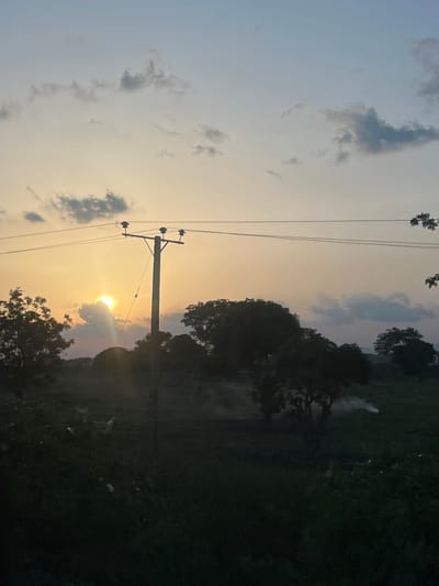 The image portrays a rural landscape in Rinjim Gani, Nigeria, captured during sunset. The sky displays a gradient from light blue at the zenith to a prominent golden-orange at the horizon. Scattered grey clouds are visible, with one cloud partially obscuring the bright, intense sun, which emits discernible rays. A wooden utility pole, equipped with three insulators, supports overhead power lines that stretch horizontally across the middle of the frame, silhouetted against the luminous sky. The midground and foreground consist of dark, heavily silhouetted trees and dense vegetation. A plume of white smoke is observed rising from the ground in the mid-right portion of the image, consistent with localized burning activity. No human figures or discernible structures other than the utility pole are present.