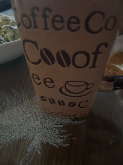 The image shows a close-up of a brown coffee mug with "Coffee Coof" and coffee bean designs. The mug is sitting on a dark brown surface, possibly a table, which has a decorative, white, fern-like placemat or coaster beneath it. To the left of the mug, a white bowl containing a light-colored, possibly creamy food item is partially visible. The background is dark and out of focus. There are no visible people or specific landmarks that would indicate a location in Rosenovo, Bulgaria.