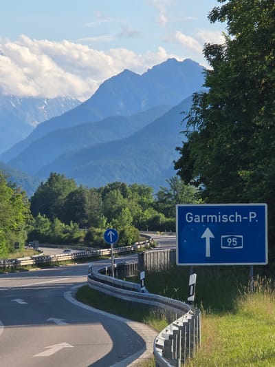 The image displays a multi-layered landscape at a highway interchange near Ohlstadt, Germany, under bright daylight with a partly cloudy sky. In the immediate foreground, a patch of sunlit tall green grass occupies the right, bordering a grey metal guardrail that curves towards the left. This guardrail, equipped with black and white reflective markers, separates the grass from an asphalt road surface. White directional arrows are painted on the asphalt, one pointing straight ahead and another indicating a slight left turn onto a merging lane.

To the right of the road and guardrail, a prominent blue rectangular road sign is positioned, displaying "Garmisch-P." in white uppercase letters, a large white upward-pointing arrow, and the number "95" enclosed in a white rectangle. This sign indicates the route to Garmisch-Partenkirchen via Bundesautobahn 95. Further along the curving road to the left, a smaller circular blue sign with a white upward arrow is visible.

The midground features a complex system of curving highway lanes, also lined with grey metal guardrails. Several vehicles, appearing as dark shapes (cars or small trucks), are discernible on these roads in the distance. Dense green foliage, comprising various deciduous trees and some conifers, flanks the roadways and fills the valleys, obscuring parts of the distant terrain. A large, leafy tree partially frames the right side of the image, with its branches extending towards the center.

In the background, a majestic mountain range dominates the horizon. These mountains exhibit a distinct blue hue due to atmospheric perspective, with closer ranges appearing darker and more distant ones progressively lighter. Patches of white snow or ice are visible on the higher peaks, particularly towards the left. The sky above is a clear light blue, adorned with scattered white, fluffy cumulus clouds, which are notably concentrated over the left section of the mountain range. The scene is illuminated by strong sunlight, casting long, defined shadows from the guardrails and trees onto the road surface, suggesting either morning or late afternoon.
