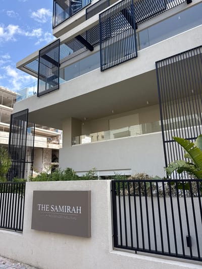 The image displays a multi-story residential building identified as "THE SAMIRAH: LUXURY RESIDENCES IN ELLINIKO," located in the Municipal Unit of Elliniko, Greece. The building's exterior is characterized by light-colored textured walls, multiple balconies equipped with glass railings, and a series of black, vertical-slat panels positioned as architectural screens or sun shades. A dark gray rectangular sign with white lettering is affixed to a low, light-colored perimeter wall in the foreground, behind which a black vertical-bar metal fence runs. Green foliage, including large-leaf plants and smaller bushes, is present behind the fence and along the building's base. To the left, a skeletal concrete structure of a building under construction or demolition is partially visible. The sky above is partly cloudy blue. No persons are present or engaged in any activity within the frame.