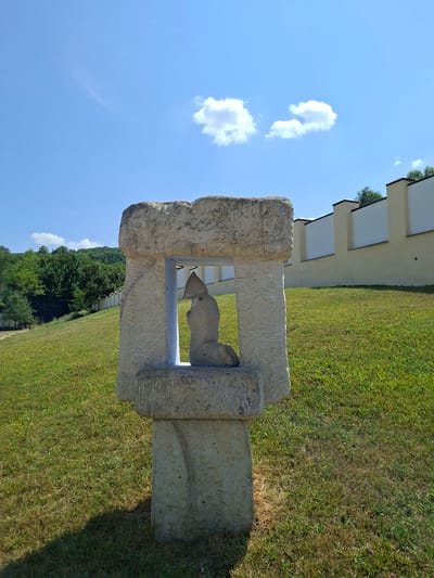 An outdoor, eye-level, medium shot shows a stone sculpture with a figure inside, located in the city of Condrița, Moldova. The sculpture is made of light-colored, rough-hewn stone, with visible scratches and carvings on its surface. The central part of the sculpture frames a smaller, abstract figure that appears to be kneeling or seated, wearing a hooded garment. This figure is also made of stone. The sculpture stands on a grassy knoll under a bright blue sky with scattered white clouds. Behind the sculpture, a light yellow wall with dark-topped vertical pillars stretches across the middle ground. The wall is set against a backdrop of lush green trees on a hillside to the left and further down the slope to the right. The grass in the foreground is a mix of green and dry patches, with some small yellow and pink wildflowers. A dark shadow is cast by the sculpture on the grass to its left. The overall scene depicts a sunny day in a rural or park-like setting.