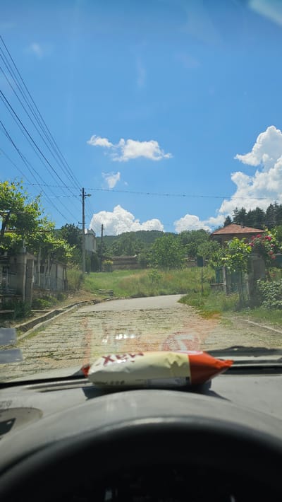 The image depicts a view from inside a vehicle, looking out onto a village road in Slatina, Bulgaria. The road is paved with cobblestones and curves to the right, leading uphill. On either side of the road are houses with red-tiled roofs and surrounding vegetation, including trees and bushes. A power line runs overhead, with poles visible. The sky is blue with scattered white clouds, indicating a sunny day. In the foreground, on the dashboard of the car, rests a snack bar. The scene is peaceful and captures a typical rural Bulgarian setting.