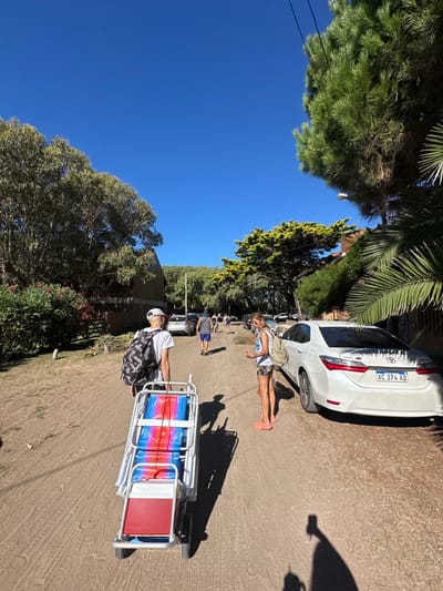 A daytime scene shows a sunny dirt road. A white car is parked. A woman in a white shirt and shorts stands near the car. A man pulls a cart carrying beach chairs. Trees border the road. The sky is blue. A shadow is cast on the road. Other people and cars are visible in the background.