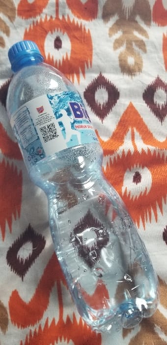 A clear plastic bottle of "BIG" brand drinking water rests horizontally on a patterned textile surface. The bottle is approximately two-thirds full of liquid and exhibits external condensation droplets. It is sealed with a blue screw cap. The label, predominantly white and blue, prominently features the word "BIG" in blue, accompanied by a white silhouette of a figure, likely a runner. Additional text on the label includes "PREMIUM DRINKING WATER," a QR code, nutritional information, and a NAFDAC logo, indicating its Nigerian origin. The background textile displays an ikat-style pattern with repeating geometric and stylized flame-like motifs in orange and dark brown hues on a white or cream base. No individuals, actions, or direct interactions are visible. The scene is depicted in a setting within the city of Ado-Ekiti, Nigeria.
