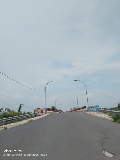 The image shows a view from the top of a bridge in Kecamatan Gabuswetan, Indonesia. It's a clear, sunny day with some clouds in the sky. The bridge is paved with gray asphalt and has a metal guardrail on both sides. There are several street lights along the bridge, and some electrical wires run overhead. The bridge is going downhill from the point of view of the camera and leads to a straight road. The road is empty of vehicles except for one motorcycle at the bottom of the hill, and there are only a few people walking in the distance. The road is slightly cracked and worn. Green foliage is visible on both sides of the bridge. A cell phone's timestamp is visible in the bottom right corner. The timestamp indicates the photo was taken on 10 April 2025 at 10:52. The location is identified by coordinates (-6.375, 108.055).