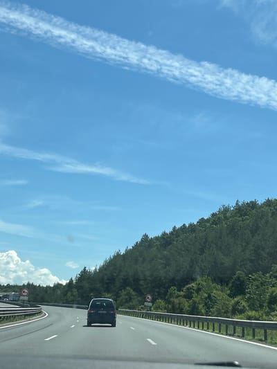 The image depicts a highway scene from the perspective of a vehicle in motion, suggesting it is taken from a car driving on the road. The road is paved and appears to be a multi-lane highway with clear lane markings. There is a guardrail running along the right side of the road. On the right side of the highway, a dense forest of green trees covers a sloping hillside. The sky is a vibrant blue with some wispy white clouds and a distinct contrail stretching across the upper portion of the frame.

In the foreground, a dark-colored minivan is visible in the right lane, traveling in the same direction as the viewpoint vehicle. The minivan has its license plate visible, though unreadable in detail. On the left side of the highway, there is a traffic sign with a red circle and a horizontal bar, indicating a restriction, possibly for trucks or certain types of vehicles, though the specific symbol within the circle is not entirely clear. Further in the distance on the left, more vehicles, including what appear to be trucks, are visible on a different section of the highway. The overall environment suggests a rural or semi-rural setting with natural vegetation bordering the highway. The specific location of "Novi han, Bulgaria" is not visually evident in the image; this information would need to be inferred or provided externally.