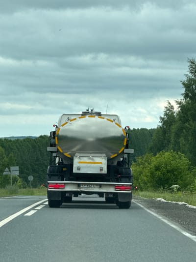 The image depicts the rear view of a silver tanker truck driving on an asphalt road in the countryside, possibly in or near Новая Казмаска, Russia. The truck has a large, cylindrical silver tank with yellow reflective stripes. It has red taillights and a license plate that reads "AP 952 116 RUS". The road has white lane markings. The surrounding environment is characterized by dense green trees and foliage, with a cloudy sky overhead. There is no visible person or interaction.