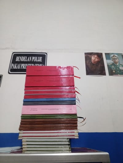 The image captures a still life composition against a white wall, possibly within a room or office. The primary subjects are a stack of books and two framed pictures. The books are of varying colors, predominantly red, pink, blue, and green, stacked vertically and suggesting they are likely notebooks or journals. Some of the books have visible ribbons or bookmarks protruding from them, indicating they are in use or have been recently accessed.

The framed pictures depict two individuals. The picture on the left shows a young man with dark hair, possibly an actor or musician, captured in a close-up portrait. The picture on the right features a man in a military uniform, wearing a cap and adorned with medals, suggesting a formal or official context.

Visible text on the wall includes a black and white sign with the words "BENDELAN POLIJIE" and "PAI PRINTED SEMUA" in white lettering. This text, combined with the military imagery, might imply a connection to law enforcement or a specific organizational context. The date "2016" is also visible on one of the books.

The lighting appears to be artificial, casting no discernible shadows that would indicate a specific time of day. The background wall is plain white, with a blue stripe running horizontally across the lower portion of the frame, possibly indicating a painted partition or furniture. The overall scene evokes a sense of order and documentation, with the stacked books suggesting a collection of records or personal writings. The location context provided is Jember, East Java.