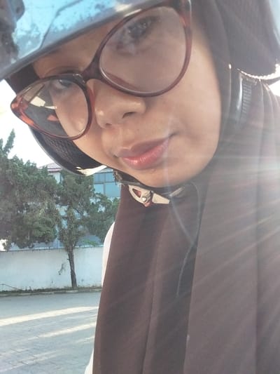 A close-up, selfie-style shot shows a person wearing a helmet and glasses. The individual's face is partially obscured by the helmet and the angle of the photo. The person has on glasses with reddish-brown frames and appears to be wearing a brown hijab or scarf. The background shows trees and a wall, suggesting an outdoor urban environment, possibly in Macege, Indonesia, though no specific landmarks confirm this location. The lighting indicates it is daytime, with sun glare visible on the right side of the image. The overall scene is informal and appears to be taken while the person is on the move, possibly on a motorcycle.