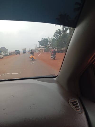 This is a photograph taken from inside a vehicle looking out through the windshield at a road scene. The road is paved with a reddish-brown surface, characteristic of Imilike, Nigeria. The sky is overcast and hazy, suggesting a potentially humid or dusty atmosphere common in the region.

In the foreground, the interior of the car is visible, including the dashboard and the windshield, which has a visible crack running across it. The perspective suggests the vehicle is moving.

On the road, several vehicles and people are present. Two individuals are riding motorcycles. The motorcycle closer to the foreground has a person with a dark patterned shirt and a pink backpack, riding it. The motorcycle further back has a person with a dark shirt carrying two large yellow containers on the back.

In the distance, there are other vehicles, including trucks. Buildings line the side of the road, suggesting a settlement or town. Palm trees and other lush green vegetation are visible in the background, consistent with the tropical environment of Nigeria. The overall scene depicts a busy, yet somewhat hazy, road in a Nigerian town.