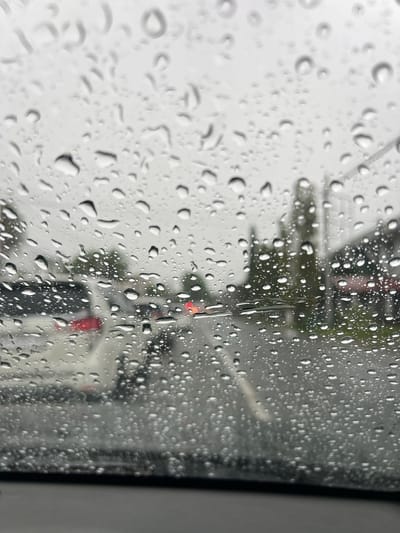 This is a photograph taken from inside a vehicle, looking through a rain-streaked windshield. The scene depicts a street in Lampisang, Indonesia, during a downpour. The primary subjects are vehicles on the road, including a white car with a red taillight visible in the left portion of the frame, and other cars further down the road, their headlights blurred by the rain. Trees and buildings line the street, but their details are obscured by the water droplets on the glass and the general overcast conditions. The overall atmosphere is one of a dreary, wet day, with visibility reduced due to the weather.