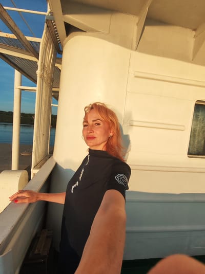 A woman with ginger hair and red lipstick poses for a selfie on a boat's deck, with the city of Bol'shoi Dvor, Russia, in the background. She is wearing a black, high-necked top with white lettering and a circular emblem on the left sleeve. Her left hand rests on the boat's railing, and her right arm extends towards the camera. The background shows a body of water, a dock, and a treeline under a clear blue sky, bathed in the warm glow of the setting sun. The boat's exterior is painted white, with a large cylindrical structure and windows visible.