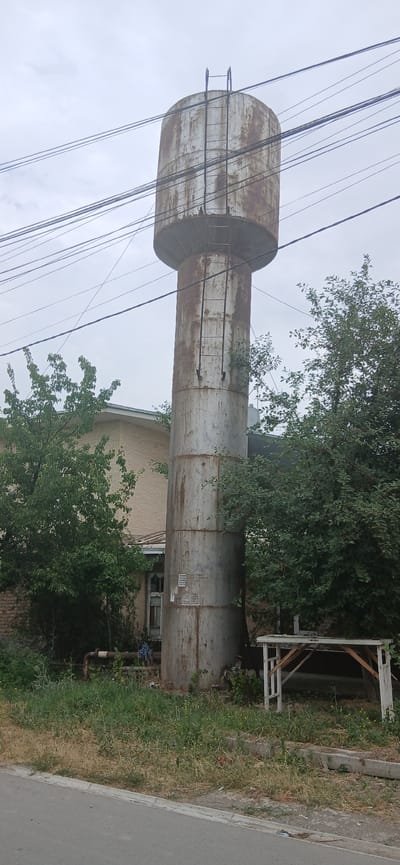 This media file captures a tall, cylindrical, weathered water tower, likely made of metal, with visible signs of rust and peeling paint. The tower is oriented vertically, suggesting the photo was taken at an angle. Electrical wires crisscross the sky and the tower, with some appearing to be attached to or supported by the structure. A metal ladder runs up the side of the tower, indicating it is accessible.
The water tower is situated in an outdoor setting with overgrown vegetation, including trees with dense green foliage, growing around its base and partially obscuring it. Behind the tower, a brick building with windows is visible, suggesting an industrial or utility area. To the right, a paved area, possibly a road or sidewalk, runs alongside a strip of dry, overgrown grass and weeds, with a simple, dilapidated wooden structure in the background.
The sky is overcast with diffused light, indicating a cloudy day. The overall atmosphere is somewhat utilitarian and perhaps neglected, given the state of the water tower and the surrounding vegetation. There is no clear activity or event taking place in the image; it appears to be a static scene.
On the side of the water tower, some white markings are visible. They appear to be stenciled numbers or symbols, arranged in a rectangular pattern, but the text is too blurry to be deciphered. The location context provided is Andijani, Uzbekistan.