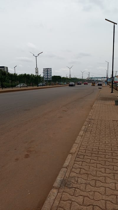 The image depicts a street scene in Kawo, Nigeria. The view is looking down a wide asphalt road with sparse traffic, including cars and a yellow auto rickshaw. A patterned brick sidewalk runs along the right side of the road. Tall streetlights line both sides of the street. A large billboard stands on the left side of the road, behind a green metal fence and some vegetation. In the distance, a sign appears to read "KAWO FOR REPS". The sky is overcast. A few people are walking on the right side of the road in the distance.