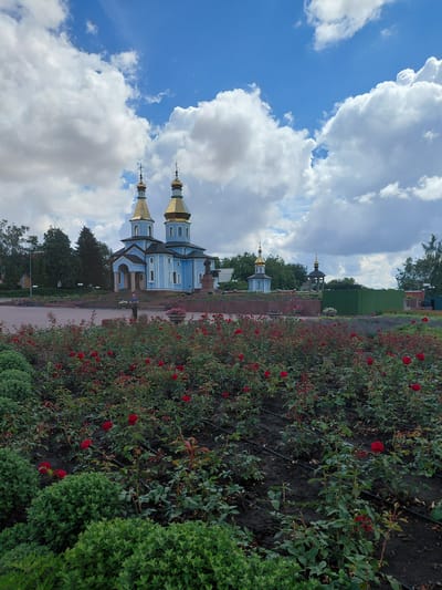 In the city of Kovalivka, Ukraine, a tranquil scene unfolds. A prominent blue church, adorned with golden domes, stands majestically against a backdrop of a dynamic sky, its facade illuminated by the natural light. In front of the church, a meticulously maintained garden bursts with vibrant red roses, their petals a stark contrast to the earthy soil. The garden is further enhanced by manicured green bushes, adding a layer of lushness to the landscape. A lone individual is captured in a moment of quiet contemplation, standing amidst the floral display. The overall atmosphere is one of peace and serenity, with the architectural grandeur of the church harmonizing with the natural beauty of the garden.