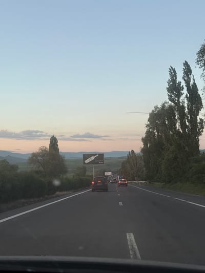 The image shows a road scene at dusk in Armenia. The perspective is from inside a vehicle, looking forward along a multi-lane highway. Traffic is light, with several cars visible ahead. The sky is a soft gradient of pale blue to pink, indicating the sunset. To the right of the road, tall, slender trees line the embankment, their leaves a dark green against the fading light. In the distance, rolling hills are visible, with a billboard featuring an advertisement for a bottled beverage on the left side of the highway. The road surface is dark asphalt with white lane markings. The overall impression is a calm, transitional moment of day.