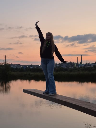 This is a vertical, medium-shot, candid photograph taken at dusk. A young woman with long blonde hair, wearing a dark hoodie and light blue jeans, is captured mid-pose on a wooden plank. Her arms are outstretched, one hand extended forward, the other pointing backward, giving the impression of flight or a superhero pose. She is looking towards the camera with a smile. The plank is positioned over a body of water, which reflects the soft hues of the sunset and the surrounding landscape.
The background depicts a serene scene with a dusky sky transitioning from light grey to soft orange and pink hues. Scattered clouds are visible. Along the far bank of the water, there is a dark line of trees and vegetation, beyond which a village or small town can be seen with houses and lights. Two tall industrial chimneys are prominent in the distance. The water's surface is calm, with gentle ripples.
The time of day is clearly evening, as indicated by the sunset colors in the sky and the reflections on the water. The weather appears calm. The location is identified as Sars, Russia. The overall mood is playful and adventurous, with the young woman expressing joy and freedom in her pose against the backdrop of a peaceful natural setting. There is no visible text in the image.
