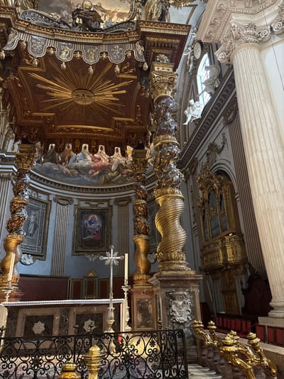 The image displays the highly ornate interior of the Cathedral of Saint Felician (Duomo di San Feliciano) in Foligno, Italy, specifically focusing on the high altar area.
A grand baldachin (canopy) dominates the central view, supported by four columns. The visible columns are dark-toned, possibly bronze, heavily sculpted with gilded spiral leaf and vine motifs, transitioning to gilded spiral fluting at their bases, and topped with elaborate gilded capitals. The baldachin's underside is dark red with a prominent central gilded sunburst motif. Its upper structure features extensive gold ornamentation and sculptural elements.
Beneath the baldachin, a rectangular altar table constructed of dark, veined marble with white decorative panels is present. Centered on the altar is a tall, silver-colored crucifix on a matching stand. To its right, a single lit white candle rests in a silver-colored candlestick. Behind the altar, the wall features vertical light-colored pilasters. Two framed paintings hang on this wall: a central one depicting the Madonna and Child with additional figures, and a smaller one to the left showing a single standing figure. Above these paintings, a semi-circular mural depicts numerous robed figures amidst clouds.
To the far right, a large, fluted white marble column with an ornate Corinthian capital ascends. Above and to its left, an arched window allows natural light into the space, flanked by white marble sculptures, including a seated figure in flowing drapery. Below the window and column, a large, gilded pipe organ is mounted on an ornate balcony. Partially visible below the organ are red upholstered pews or seating and a gilded railing.
In the immediate foreground, a black metal altar railing with intricate floral and scrollwork designs partially obscures a patterned marble floor. No people are visible within the scene.