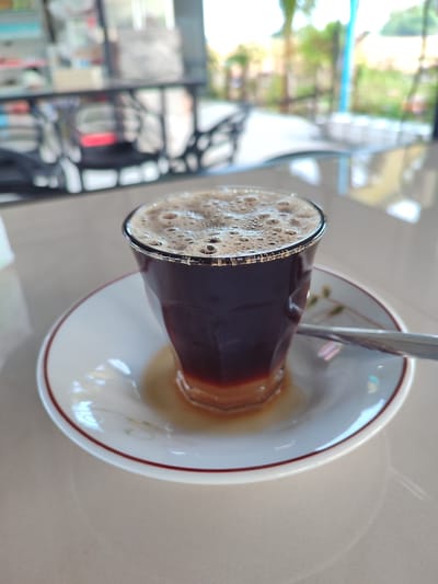 A close-up shot captures a glass of coffee with a frothy top, placed on a white saucer with a subtle floral pattern and a thin red rim. A silver spoon rests on the right side of the saucer. The coffee appears dark brown with a lighter, amber liquid pooling around the bottom of the glass. The scene is set on a smooth, light-colored surface, possibly a table, with a blurred background indicating an indoor or covered outdoor setting with seating and greenery visible. The image is taken from a slightly elevated angle, focusing on the beverage. The location is identified as Cotglumpang, Indonesia.
