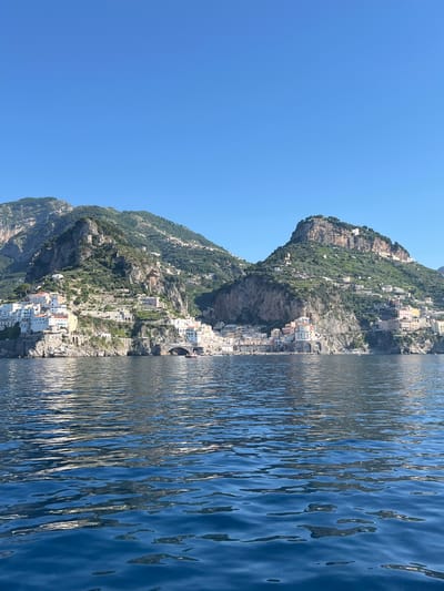 This is a horizontal, eye-level, medium-wide shot of the Amalfi Coast, specifically depicting the village of Atrani nestled at the base of steep, green mountains that rise behind it. The village is characterized by a dense collection of white and pastel-colored buildings with terracotta roofs, clinging to the rocky slopes. A notable feature is a large arched stone bridge spanning a section of the coastline. The water in the foreground is a deep blue with ripples reflecting the sunlight and the colors of the sky and land. Two small boats are visible near the shore, one of which appears to be a red dinghy. The sky is a clear, bright blue with no clouds. The image captures the architectural beauty of the Italian coast and its dramatic natural landscape. The scene is devoid of any identifiable people.
