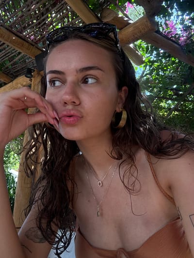 A female individual is depicted in a close-up, chest-up view, positioned outdoors under a wooden pergola adorned with natural foliage and pink-purple flowers, consistent with an outdoor setting in Lambi, Greece. The subject has dark, wet-appearing hair and wears dark sunglasses resting on her head. She is adorned with large, gold-colored hoop earrings, a light-colored ring on her left ring finger, and two layered necklaces; one features a clear, rectangular pendant, and the other a smaller, tear-drop shaped pendant. Her attire includes a bronze-colored, ribbed top. Her left hand is raised, with fingers curled and thumb resting near her chin, as her index finger touches her puckered lips, indicating a gestural expression. Her gaze is directed towards the right side of the frame. A faint, cloud-like tattoo is visible on her left upper arm. The background consists of bright green foliage with sunlight filtering through, and a black light fixture is visible on a wooden beam within the overhead structure.