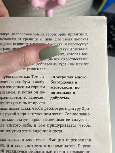 The image shows a close-up of an open book with Russian text. A person's hand with light green nail polish is resting on the left page, obscuring some of the text. The background is blurred and appears to be indoors, with hints of light-colored furniture. The text discusses a location in Argentina near the border with Chile, a high mountain peak, and a statue of Christ the Redeemer. There are no discernible elements in the image that indicate the city of Dachnoe, Russia.