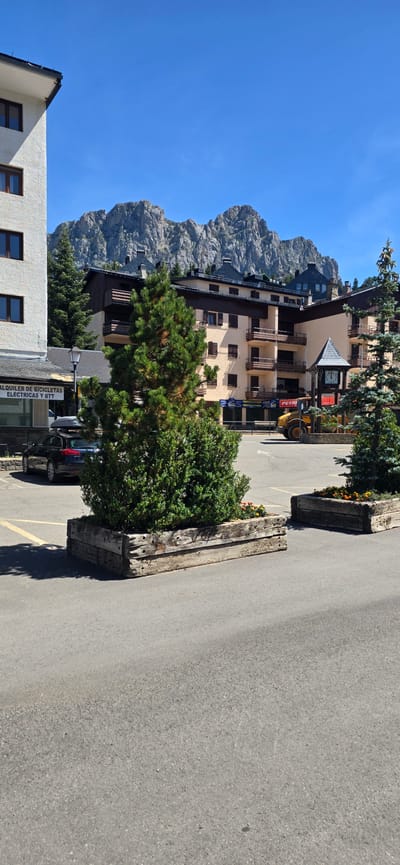 The image shows a view of the town of Sallent de Gállego, Spain, with a focus on the buildings and mountains in the background. In the foreground, there is a paved area with two large wooden planters containing green shrubs and some flowers. A dark colored car with a roof rack is parked on the left side of the frame. Behind the planters, there are several multi-story buildings. The building on the far left is white with dark windows. The central building is light beige with dark brown trim and balconies. To the right of this building is a clock tower. Further in the background, a yellow construction vehicle, likely a loader, is visible. The scene is set against a backdrop of rugged, rocky mountains under a clear blue sky. The overall impression is a sunny day in a mountainous town.