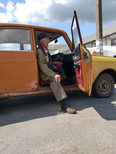 An elderly man in a light brown suit jacket, beige striped pants, a white shirt with a red and white striped scarf, and a dark baseball cap is sitting in the driver's seat of an old orange and yellow car with the driver's side door open. He is looking to his right, out of the frame. The car has visible rust and wear, particularly on the lower body panels and the wheel rim. The interior of the car shows red door paneling and black dashboard elements. The car is parked on an asphalt surface, with shadows cast by the vehicle and a concrete utility pole to the right. In the background, there are low-rise buildings with corrugated metal roofs and some advertisements on the utility pole. The sky is partly cloudy. This scene appears to be a street scene in Chudniv, Ukraine.