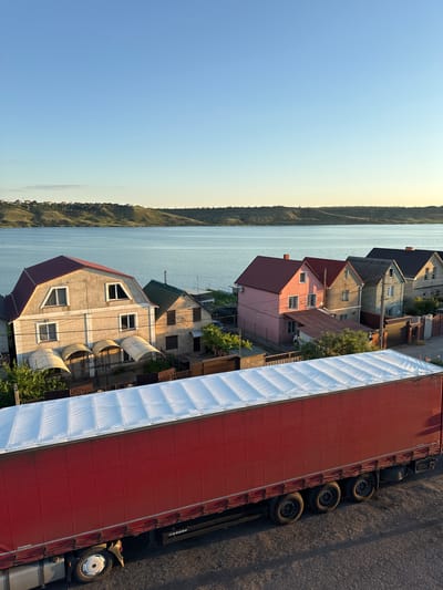 The image captures a waterfront scene in Yelyzavetivka, Ukraine. In the foreground, a large red semi-trailer truck with a white tarp-covered roof is parked on a gravel surface. The truck has multiple wheels and visible "KRONE" branding. Beyond the truck, a row of houses with distinct colored roofs (red, green, pink, gray, and dark gray) are situated along a street. The houses vary in architectural style and some have awnings over their entrances. A body of water, likely a river or lake, is visible behind the houses, reflecting the clear blue sky. The background features rolling, green hills with a sparse scattering of trees. The overall lighting suggests it is either early morning or late afternoon, with the sun casting long shadows. There are no people visible in the scene.